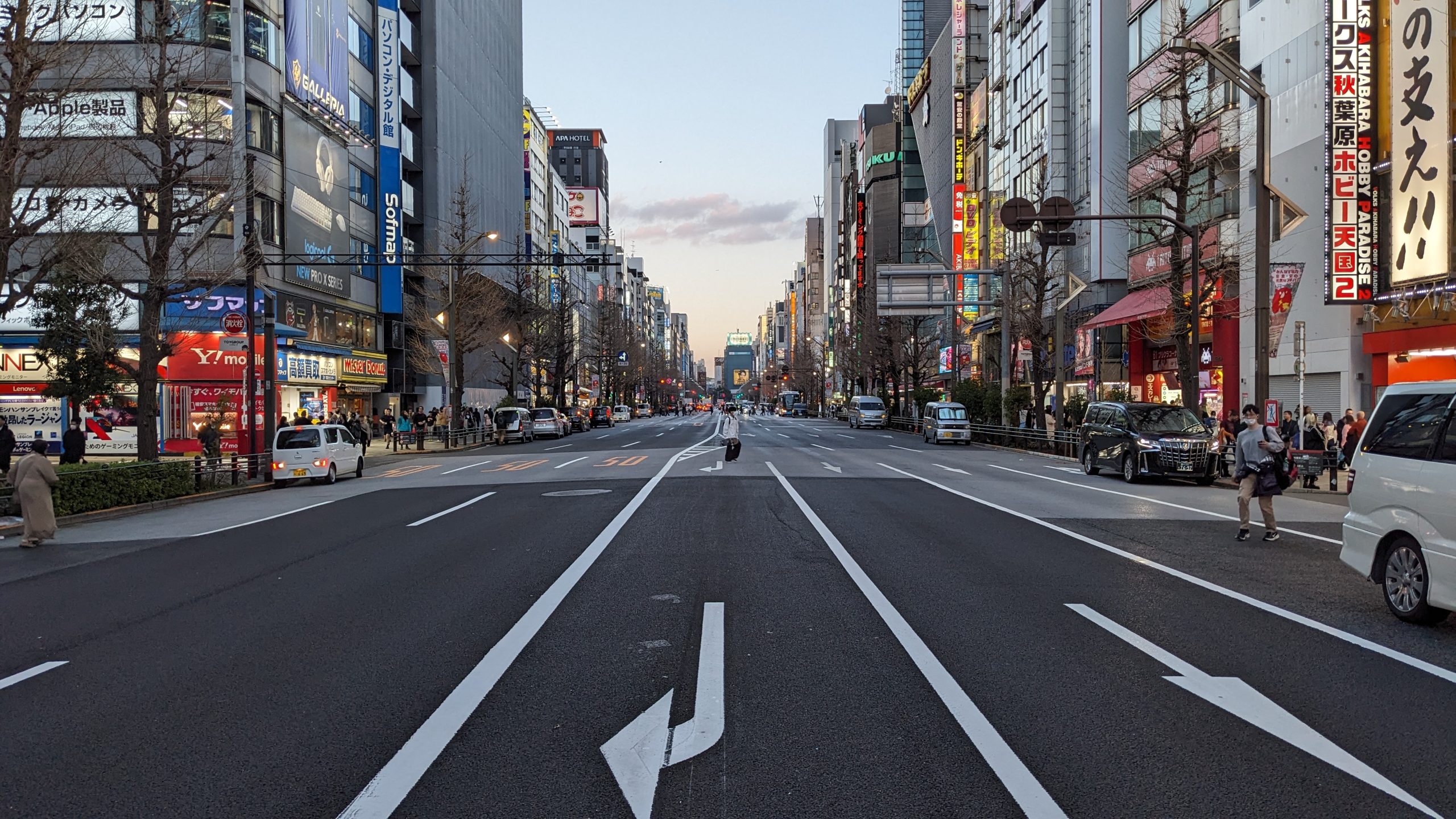Strada ampia e poco affollata nel quartiere di Akihabara a Tokyo, con segnali stradali bianchi sul manto grigio, fiancheggiata da edifici con insegne luminose e alberi spogli, e un tramonto che colora il cielo in lontananza