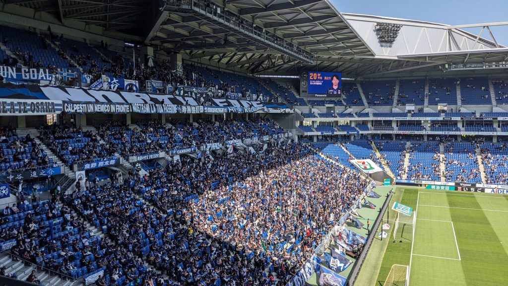 Curva piena di tifosi in un giorno di partita allo stadio di calcio del Gamba Osaka, con i fan vestiti di blu e bianco che mostrano bandiere e striscioni di tifo, sotto un cielo sereno e un'imponente struttura dello stadi