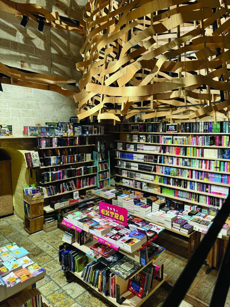 L'immagine mostra l'interno di una libreria con una grande varietà di libri esposti su scaffali e tavoli. Al centro dell'immagine si può ammirare un'installazione artistica appesa al soffitto, realizzata con strisce di legno di pioppo che creano una struttura a nastro. L'illuminazione proviene sia dall'installazione centrale che dai faretti incastonati nel soffitto di pietra, conferendo all'ambiente un'atmosfera calda e invitante. Il pavimento è rivestito di pietra, il che contribuisce all'aspetto rustico e storico del luogo.