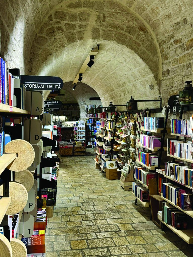 L'immagine mostra un corridoio di una libreria situata all'interno di un edificio storico con soffitti a volta in pietra. Le volte in pietra creano un'atmosfera suggestiva e tradizionale. Lungo il corridoio sono posizionati scaffali di libri etichettati con varie categorie come "STORIA-ATTUALITÀ", indicando un'organizzazione tematica. Sono visibili altri articoli, come cartoline e oggetti di cancelleria, esposti per la vendita. Il pavimento è lastricato con pietre ben adattate che rafforzano il senso di antichità del luogo. L'illuminazione è fornita da faretti moderni che contrastano con l'architettura storica, bilanciando il vecchio con il nuovo.