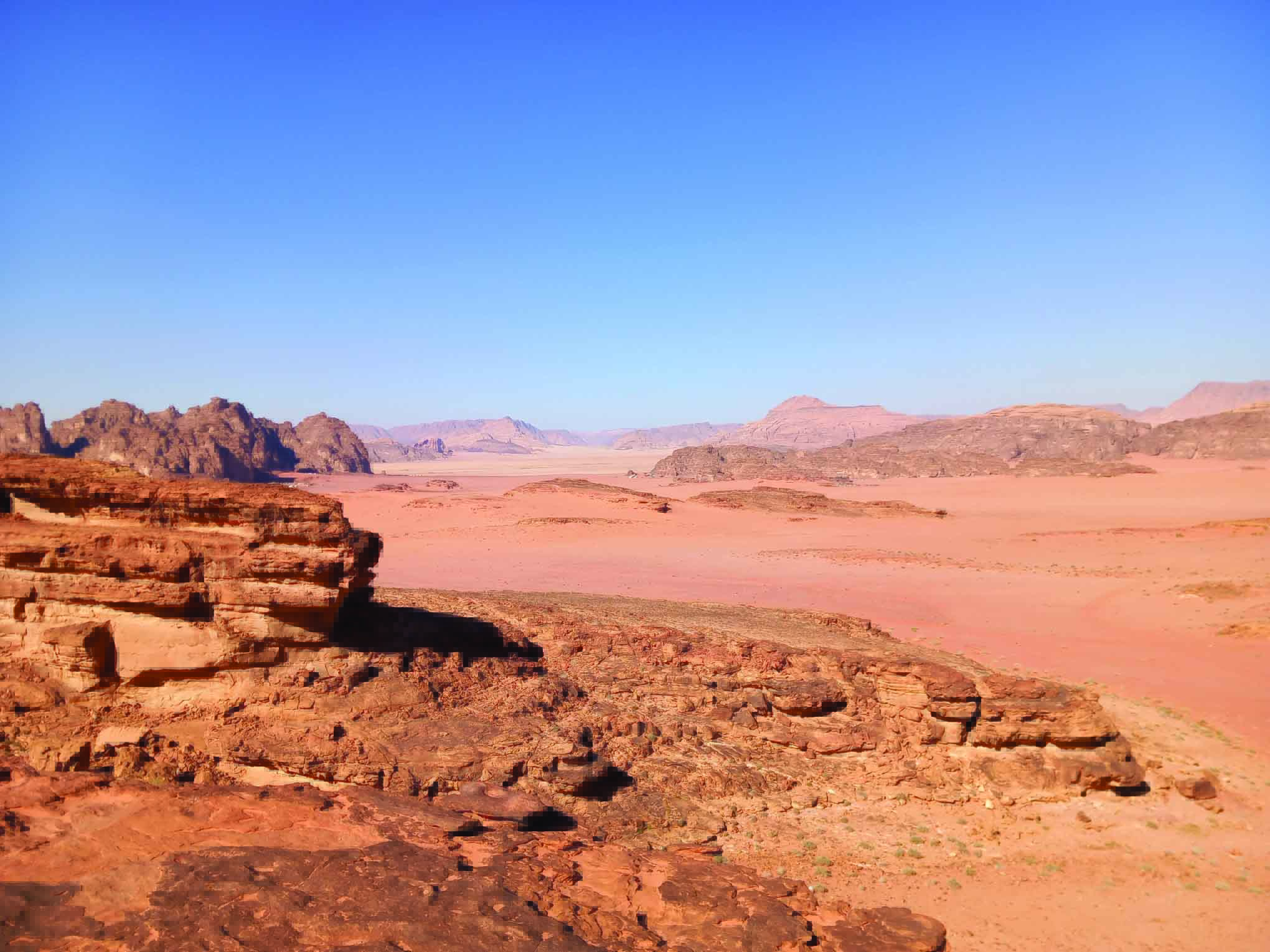 Deserto con formazioni rocciose e montagne in lontananza, sotto un cielo azzurro limpido. Ambiente arido e vasto, con sabbia e rocce rosse.