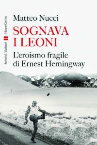 Copertina del libro "Sognava i leoni: L'eroismo fragile di Ernest Hemingway" di Matteo Nucci. La copertina mostra una persona che cammina lungo una strada di campagna innevata, calciando un oggetto in aria. Sullo sfondo ci sono colline coperte di neve e alberi. Il titolo è scritto in rosso e nero, con il logo HarperCollins a sinistra