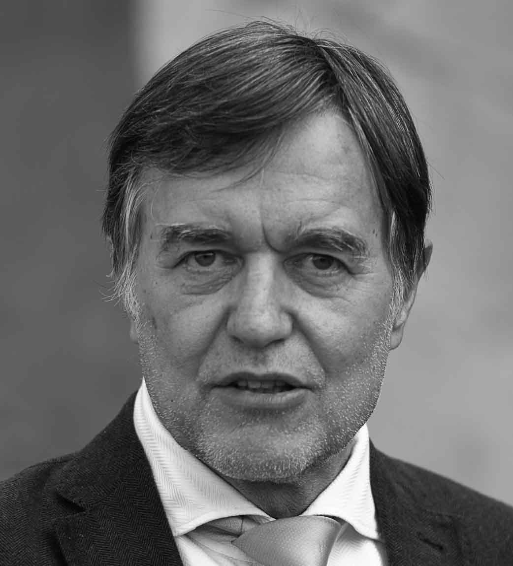 Ritratto in bianco e nero di un uomo con capelli grigi e barba, indossando un completo elegante e cravatta, con un'espressione seria e determinata.